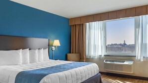 A hotel room with a large bed, a nightstand with a lamp, a view of a cityscape through the window, and an air conditioning unit. - The Capitol Hotel, Ascend Hotel Collection (Hartford)