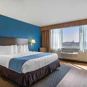 A hotel room with a large bed, a nightstand with a lamp, a view of a cityscape through the window, and an air conditioning unit. - The Capitol Hotel, Ascend Hotel Collection (Hartford)