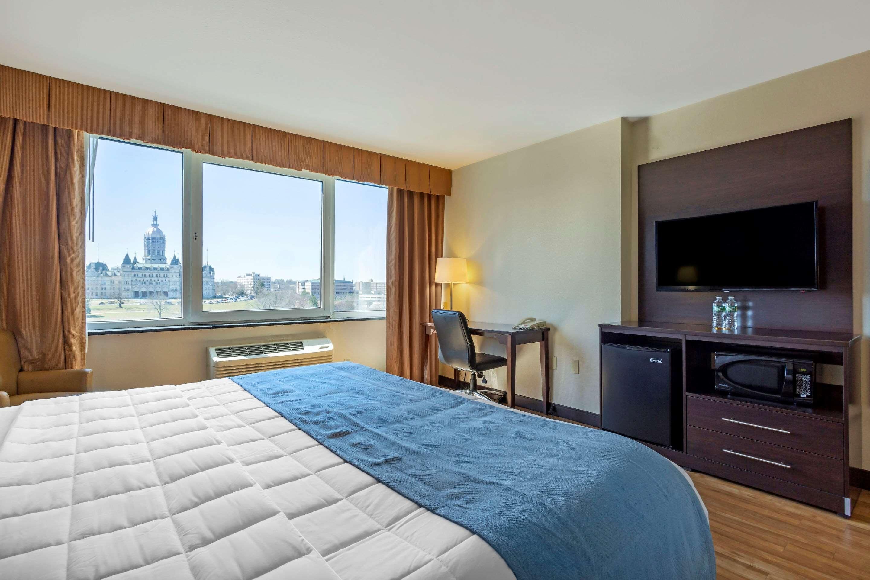 Quarto de hotel com uma cama grande, uma escrivaninha, uma televisão e vista para a paisagem urbana.