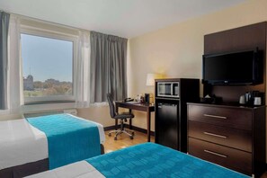 A hotel room with two beds, a desk with a computer, a microwave, a TV, and a view of a cityscape.