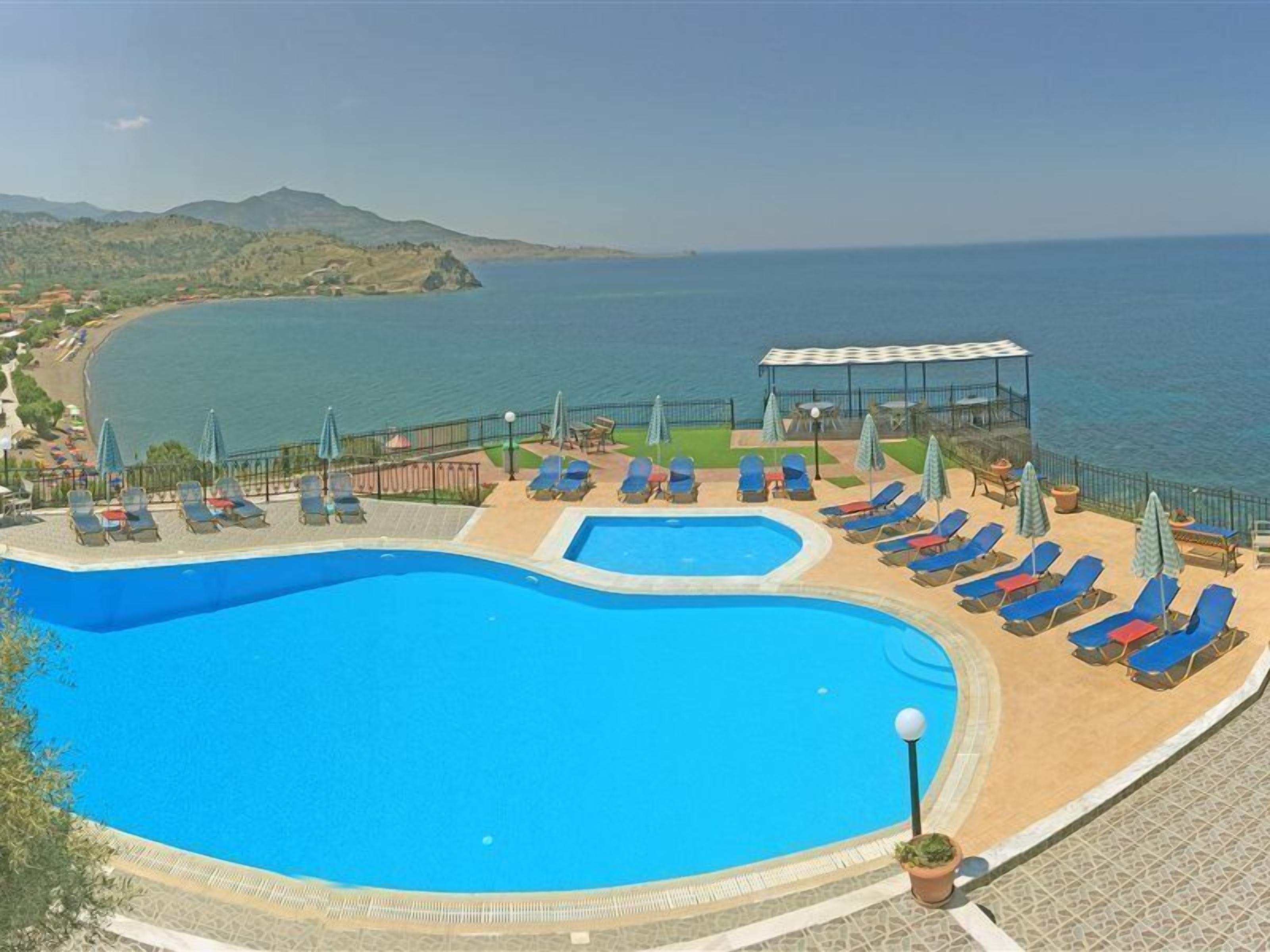 A pool area with multiple sun loungers and umbrellas, surrounded by a fence, overlooking a coastal landscape.