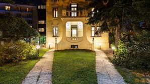 Un edificio bien iluminado y de estilo histórico, con una fachada simétrica, una entrada central y un camino pavimentado que conduce a ella.