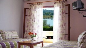 A bedroom with two beds, a wooden table, a vase with flowers, and a view of a lake through a window with floral curtains. - Gerekos Apartments (Corfu)
