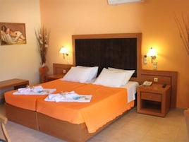 Una cama doble con colcha naranja, dos almohadas blancas, dos mesitas de noche con lámparas, una mesita con teléfono y un cuadro en la pared.