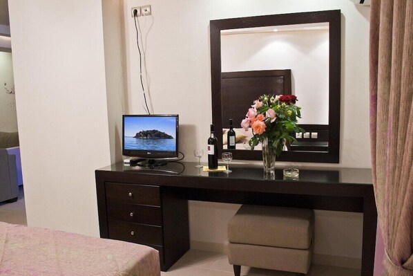 Quarto de hotel com cama, escrivaninha com computador, espelho e um vaso com flores.