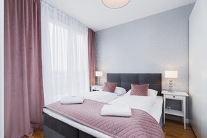 Cama doble con edredón rosa acolchado, dos mesitas de noche con lámparas, una mesita auxiliar y una ventana con cortinas de gasa.