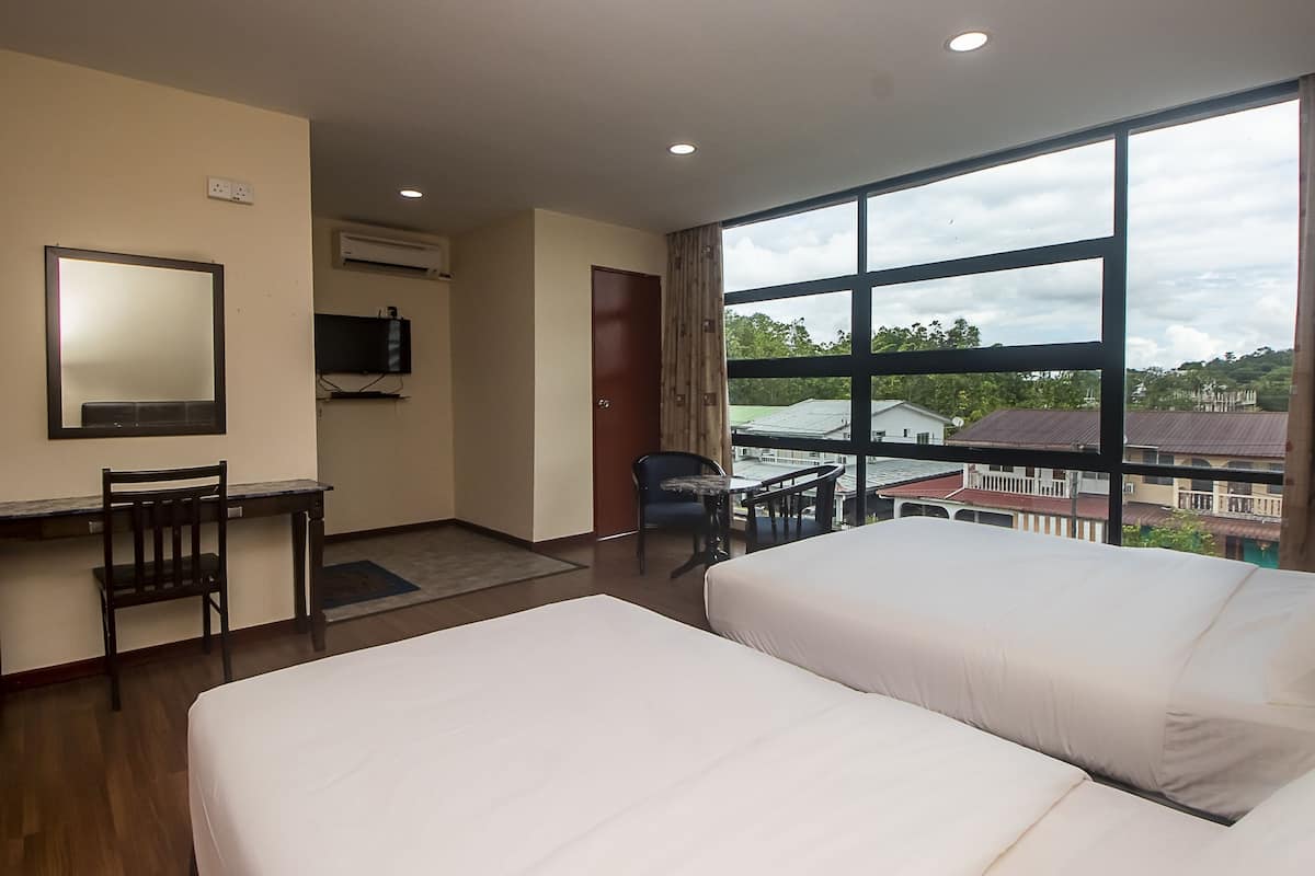 A hotel room with two beds, a desk with a chair, a TV, and a large window offering a view of buildings and trees.