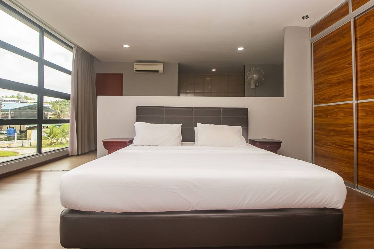 A modern hotel room with a large bed, a wooden floor, and a view of the outdoors through large windows.