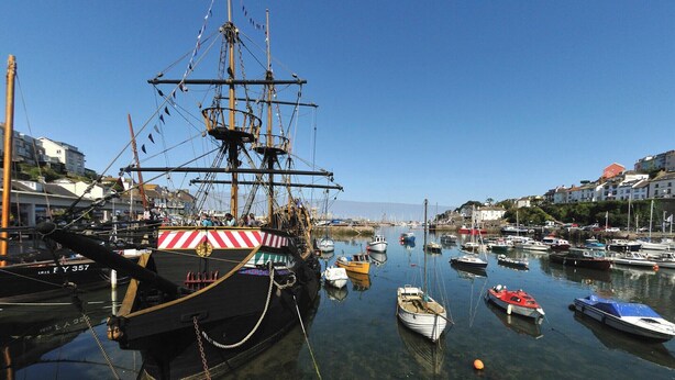 Ein Hafen mit einem großen Segelschiff, das im Mittelpunkt steht, umgeben von kleineren Booten und Gebäuden entlang des Ufers.