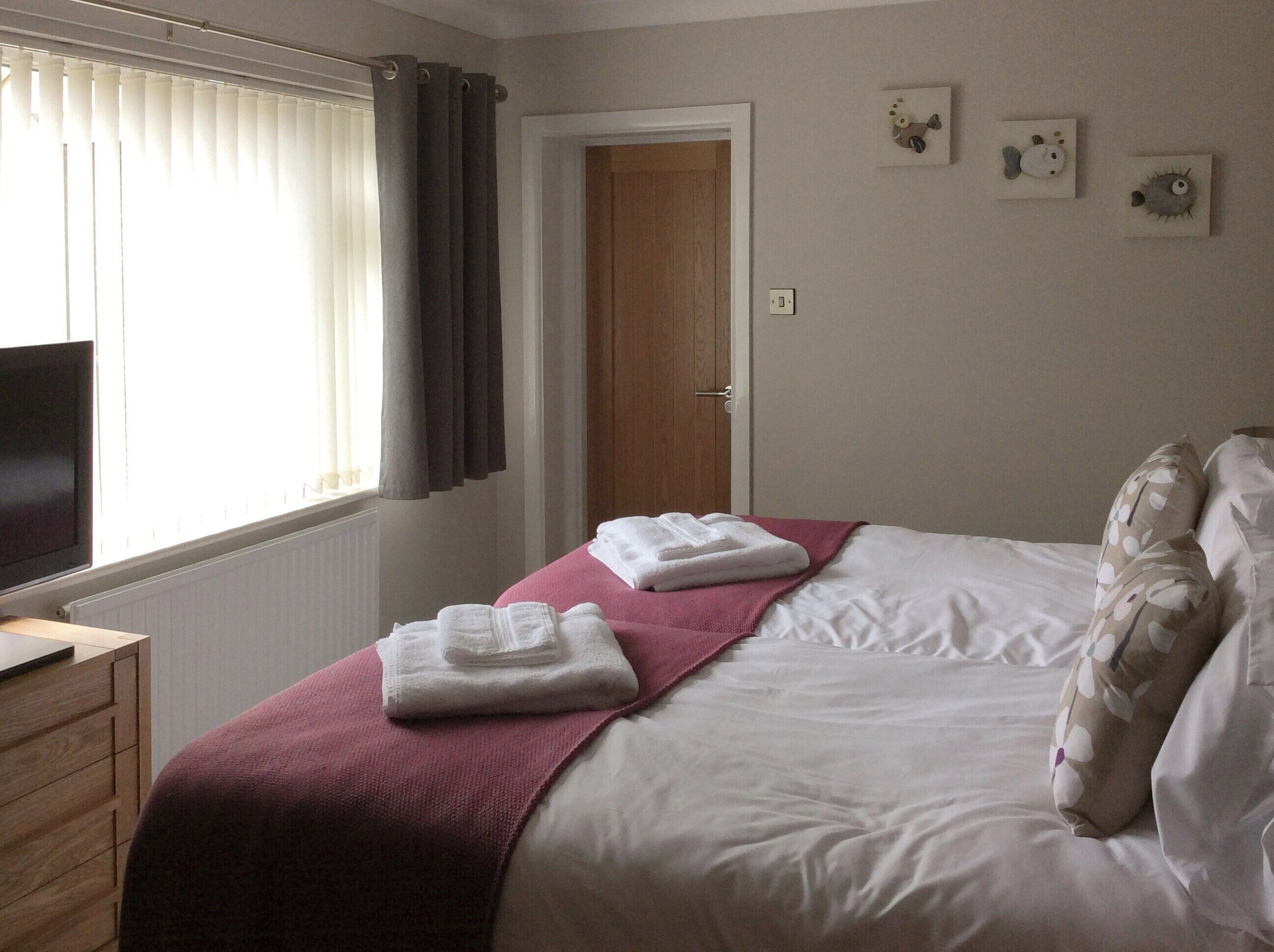 Ein ordentlich bezogenes Bett mit weißen Laken und einer rosa Decke, ein Holzkapitänsbettchen, ein Fernseher links und eine Tür mit Lichtschalter an der Wand.