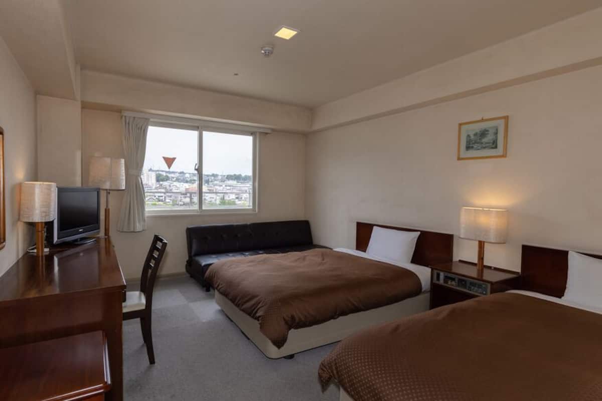 A hotel room with a bed, a desk with a television, a chair, a window with a view, and a bedside table with a lamp.
