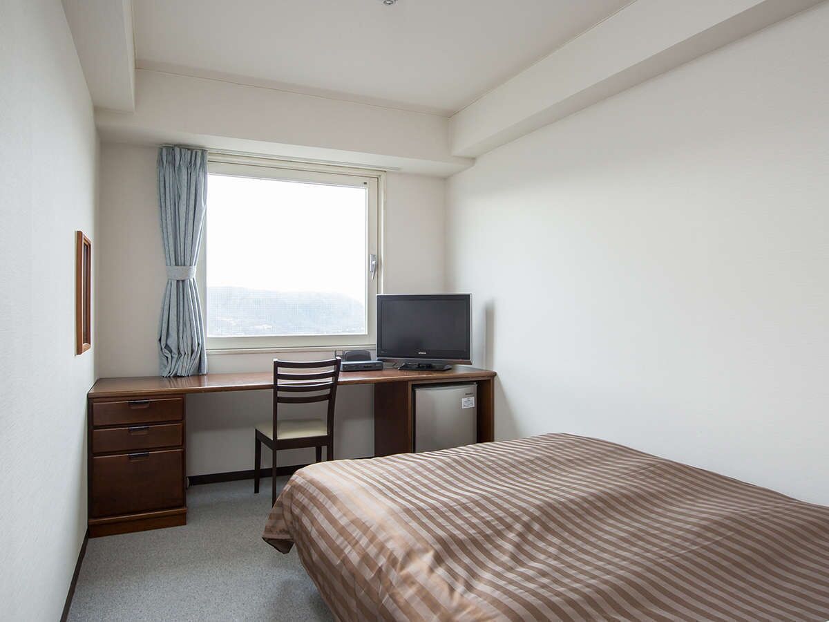 Una habitación pequeña y ordenada con una cama individual, un escritorio con un ordenador, una silla y una ventana con cortina.