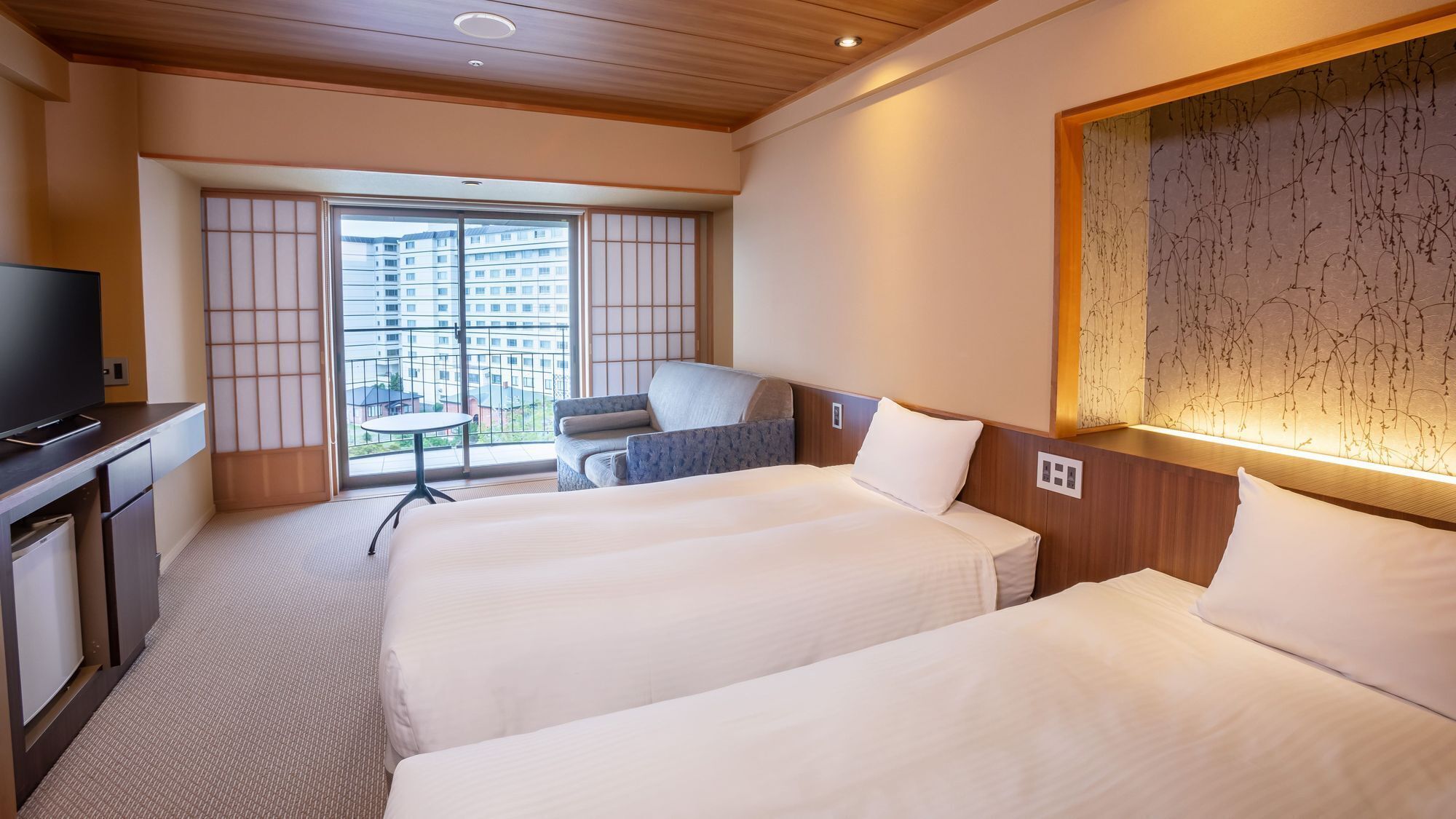 Habitación de hotel con dos camas, televisión, escritorio y vista a un edificio a través de un ventanal grande.