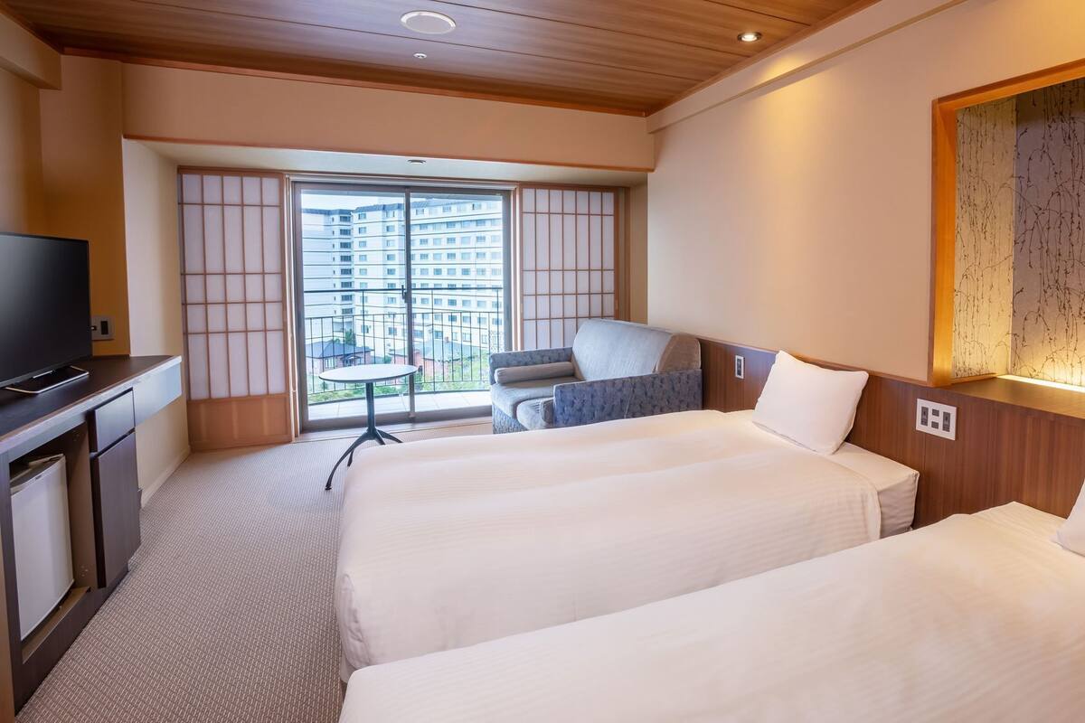 A hotel room with two beds, a TV, a desk, and a view of a building through a large window.