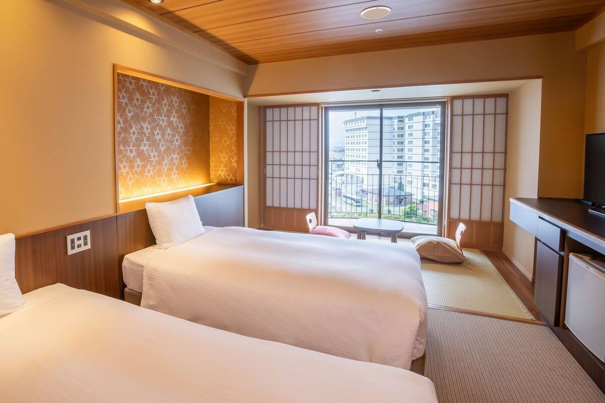 A hotel room with two beds, a TV, a balcony with a view of buildings, and a traditional Japanese sliding door.