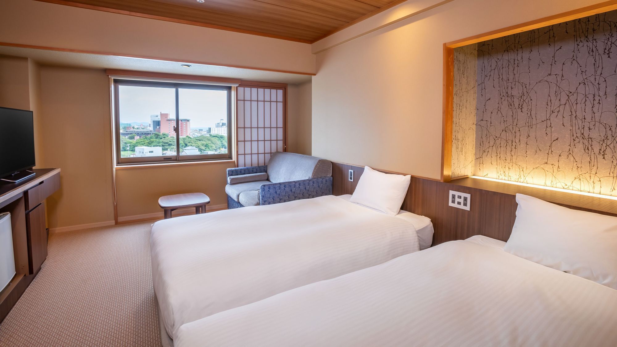 A hotel room with two beds, a small seating area, a television, and a window with a city view.