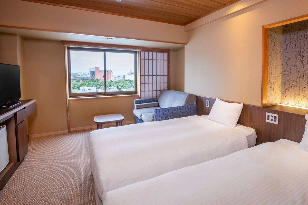 A hotel room with two beds, a small seating area, a television, and a window with a city view.