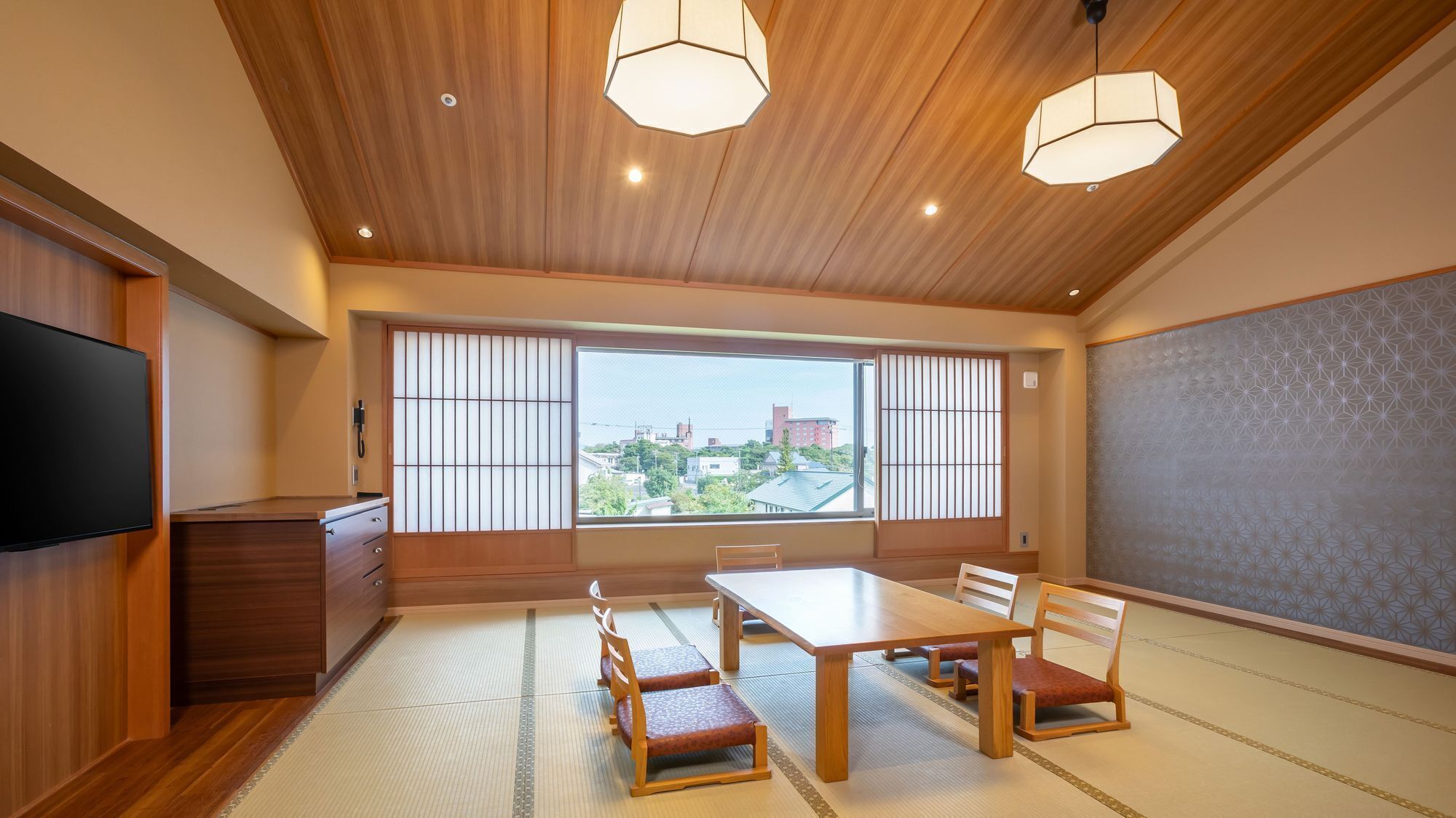 A traditional Japanese room with tatami flooring, a low table with chairs, a TV, and a large window with a view of buildings.