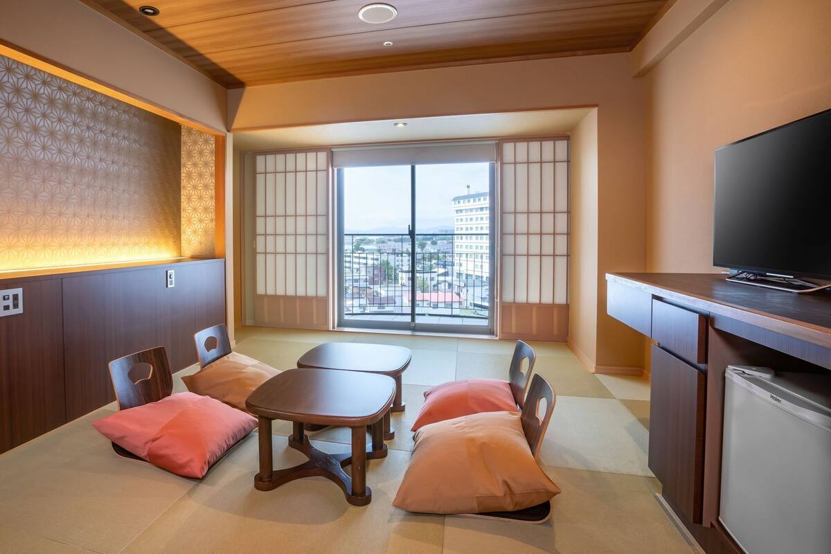 A modern hotel room with tatami flooring, a traditional sliding door, a television, and a view of the city.