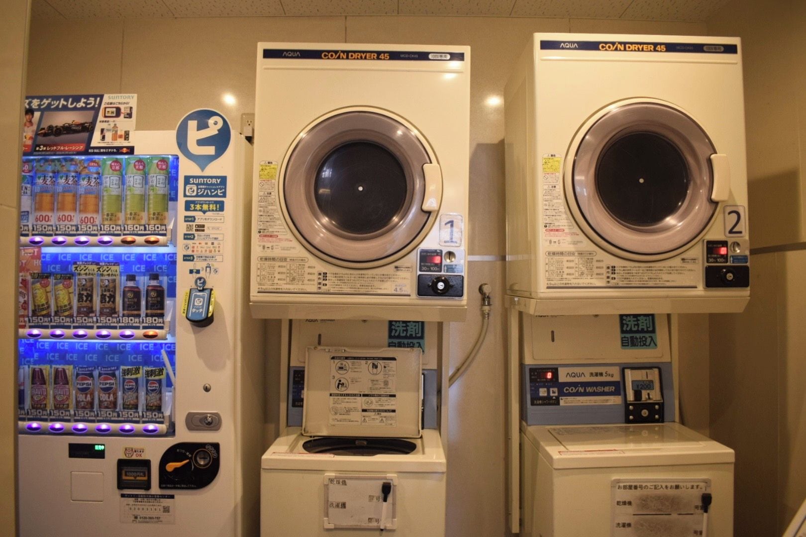 Dos lavadoras secadoras enrolladas con instrucciones en japonés, una máquina expendedora de bebidas y un pequeño cartel con un círculo azul.