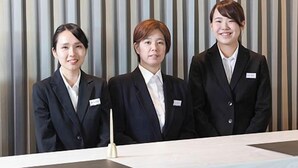 Three individuals seated at a reception desk with a vertical striped wall in the background.