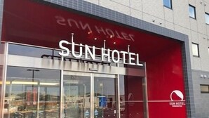 A modern hotel entrance with a red facade, large glass doors, and a visible parking area.