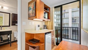 A compact kitchen with a microwave, coffee maker, and a small refrigerator. There is a wooden cabinet above the counter and a window with a view of a building.