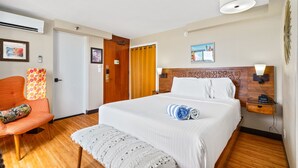 A hotel room with a large bed, a wooden floor, a chair, a bench, a nightstand, and a wall-mounted lamp.