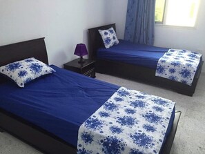 Una habitación con dos camas, cada una con una colcha azul y un cojín blanco con estampado floral azul. Hay una mesita con una lámpara morada entre las camas.