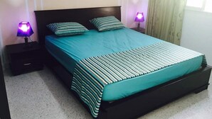 Un lit bien fait, recouvert d’une couvre-lit turquoise et agrémenté d’oreillers à rayures, avec deux lampes de chevet et une tête de lit en bois foncé.