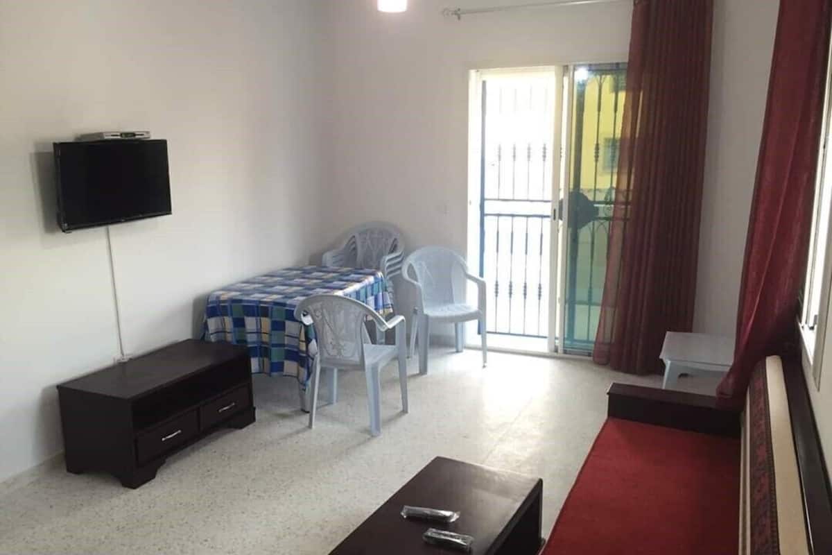 Ein Zimmer mit einem Bett, einem an der Wand befestigten Fernseher, einem kleinen Tisch, einem Sessel und einem roten Teppich.