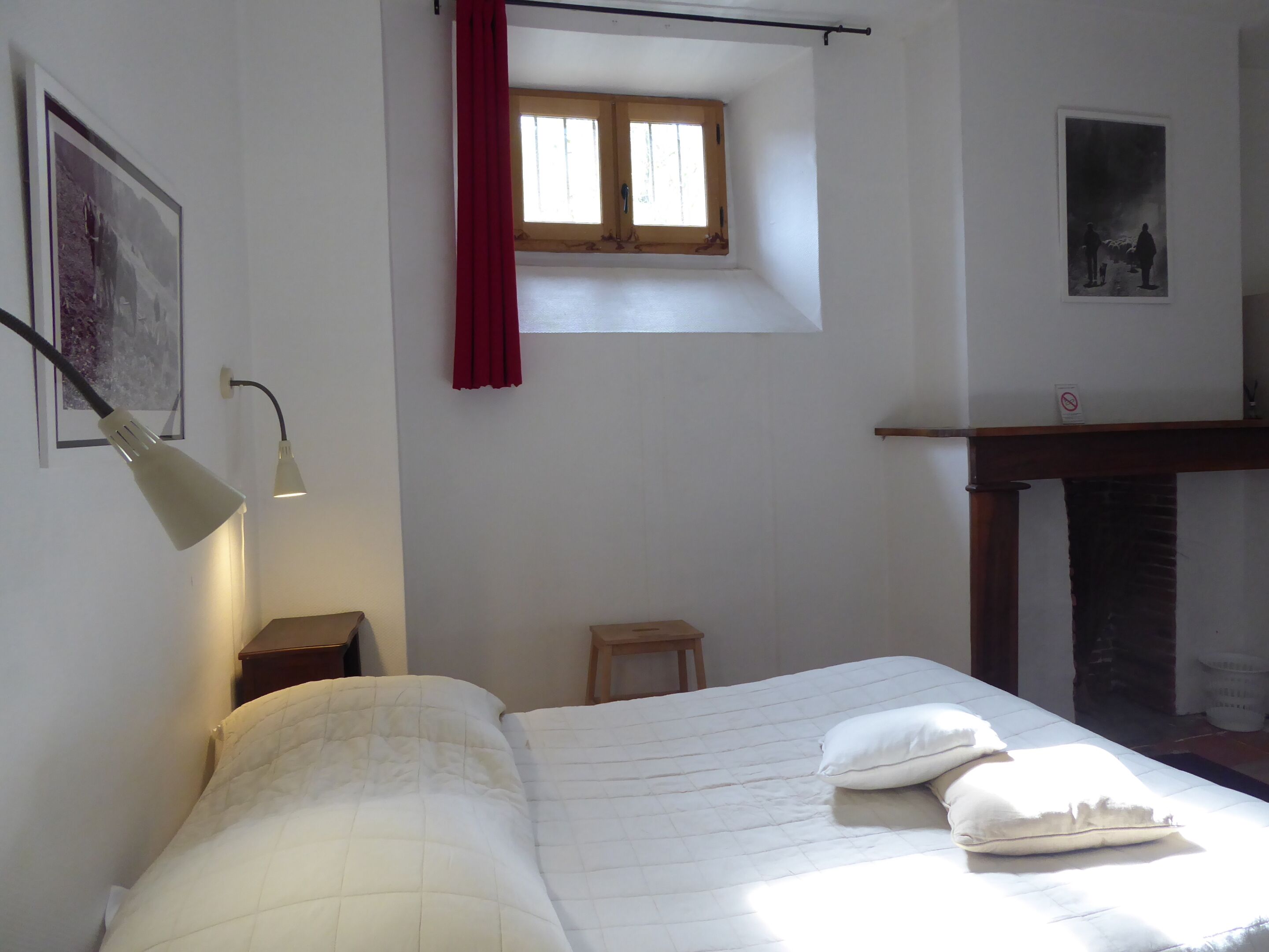 Una camera da letto con un letto, una finestra con tende rosse, una lampada a parete e un piccolo sgabello.