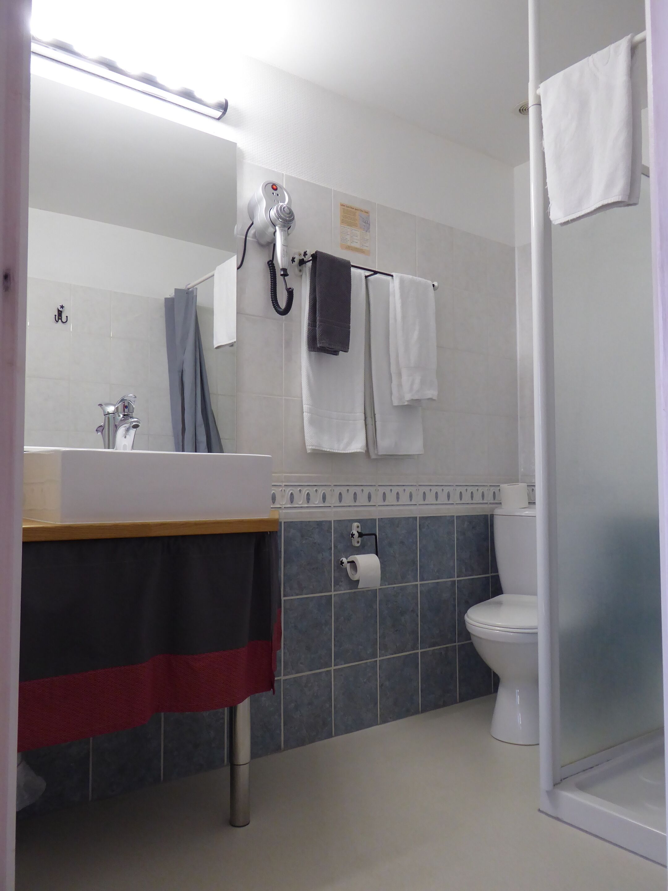Un baño con lavamanos, espejo, toallero, ducha, inodoro y un rollo de papel higiénico.