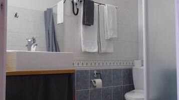 Un baño con lavamanos, espejo, toallero, ducha, inodoro y un rollo de papel higiénico.