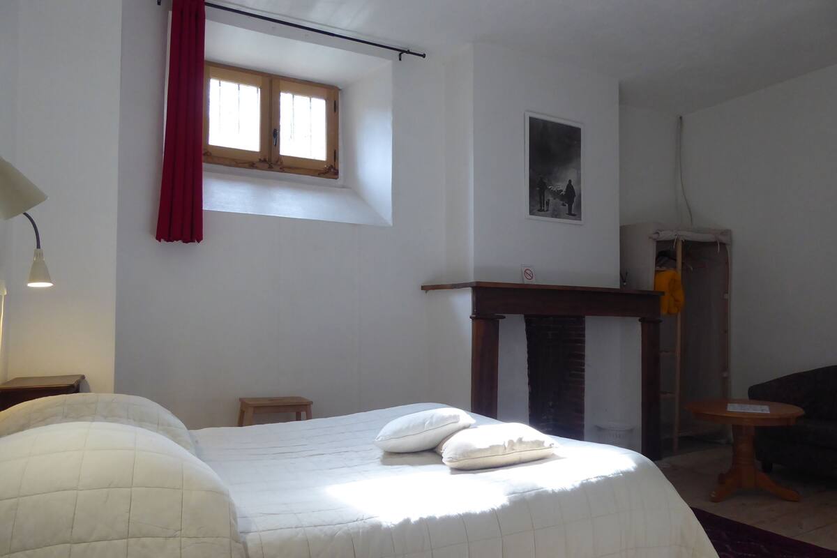A bedroom with a bed, a small table, a window with red curtains, a fireplace, and a wall-mounted picture.