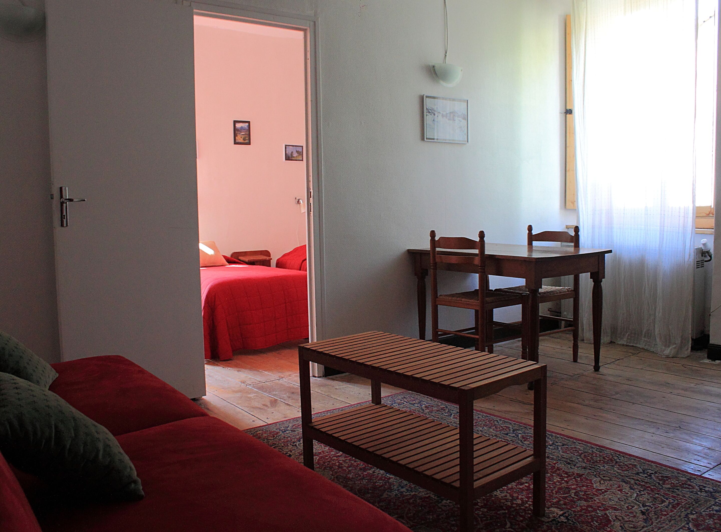 A room with a wooden dining table, two chairs, a red sofa, and a bed with red bedding.