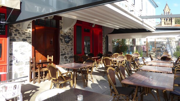 An outdoor cafe with tables and wicker chairs, stone walls, and a view of a building with a clock tower. - Auberge L'escaleta (Seix)