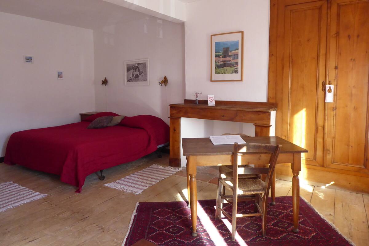 A bedroom with a bed, a wooden desk, a chair, a wardrobe, and a small table with a vase.
