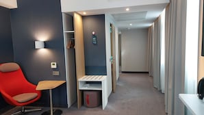 A hotel room with a red chair, a small table, a desk, a wardrobe, a mirror, and a door leading to another room.