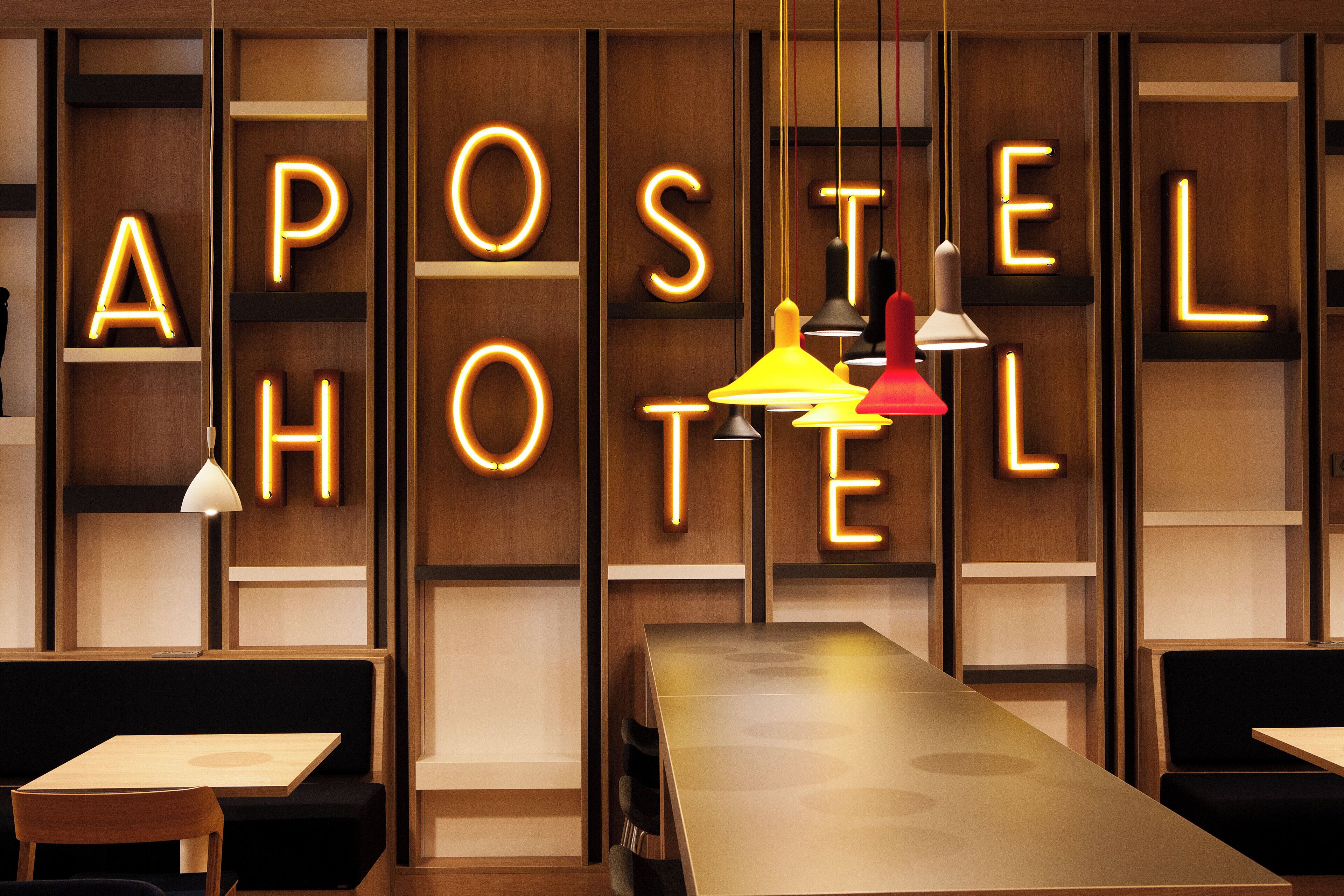 Interior de un hotel con letras iluminadas que deletrean 'APOSTEL HOTEL' en la pared, una larga mesa y asientos tapizados en negro.