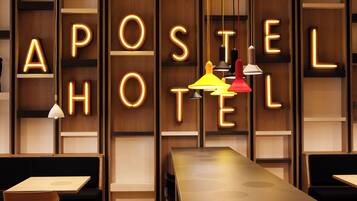 Interior de un hotel con letras iluminadas que deletrean 'APOSTEL HOTEL' en la pared, una larga mesa y asientos tapizados en negro.