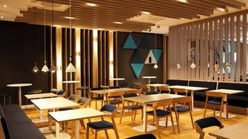 Un interior de restaurante moderno con varias mesas y sillas, decoración mural geométrica e iluminación colgante.