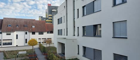 Un moderno edificio residencial con varios balcones, un camino pavimentado y un pequeño jardín con árboles.