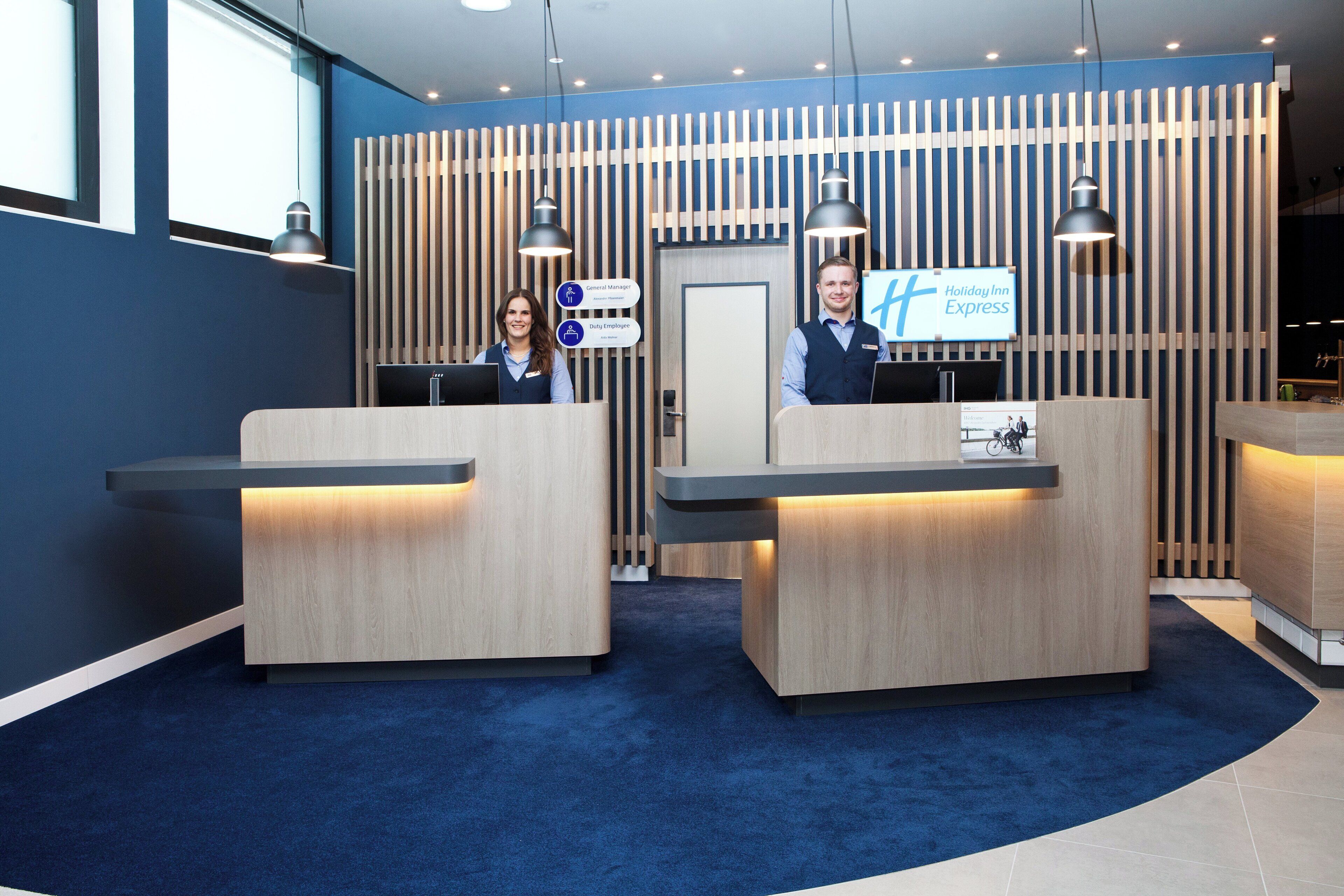 Zona de recepción con dos recepcionistas, un letrero de Holiday Inn Express y un diseño moderno con listones verticales de madera e iluminación colgante.