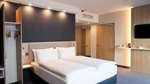 A modern hotel room with a large bed, a wardrobe, a vanity with a mirror, and a flat-screen TV mounted on the wall.