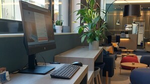 A modern office space with a long desk, computer, and ergonomic chair. There are hanging pendant lights, a potted plant, and a view of buildings outside.