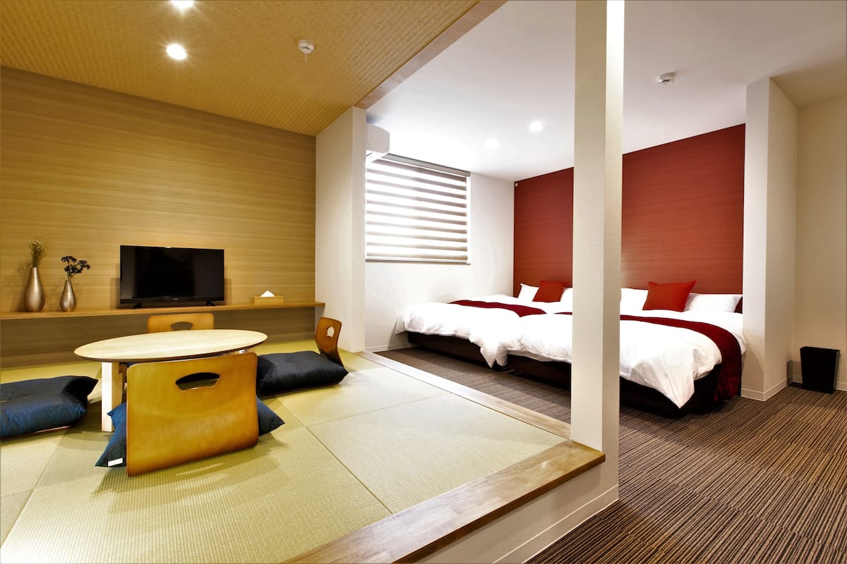 Habitación de hotel moderna con zona de estar con tatami, televisor de pantalla plana, cama con ropa de cama roja y un ventanal grande con persianas.