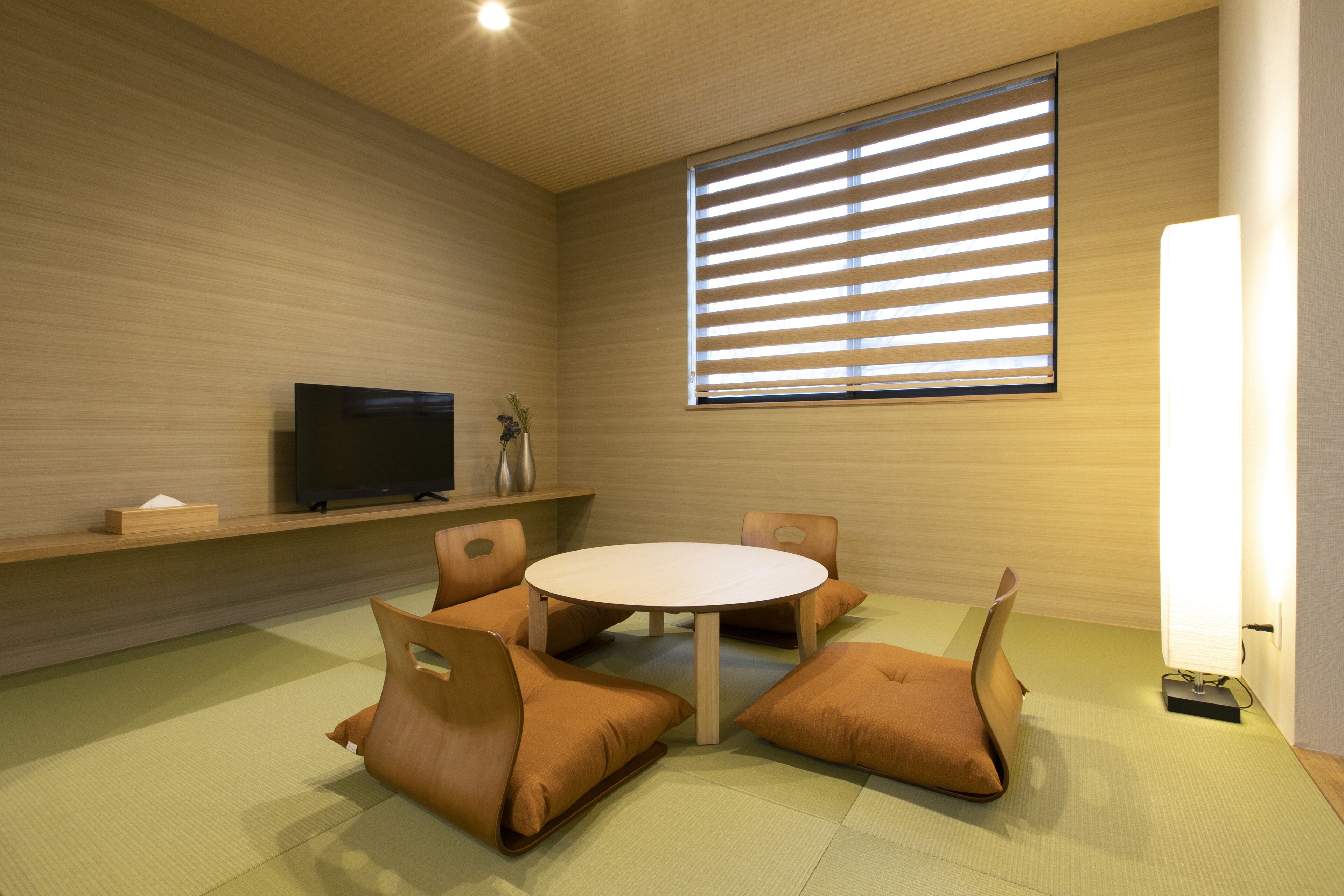 A modern room with a round table, four brown cushioned chairs, a flat-screen TV on a shelf, and a large window with blinds.