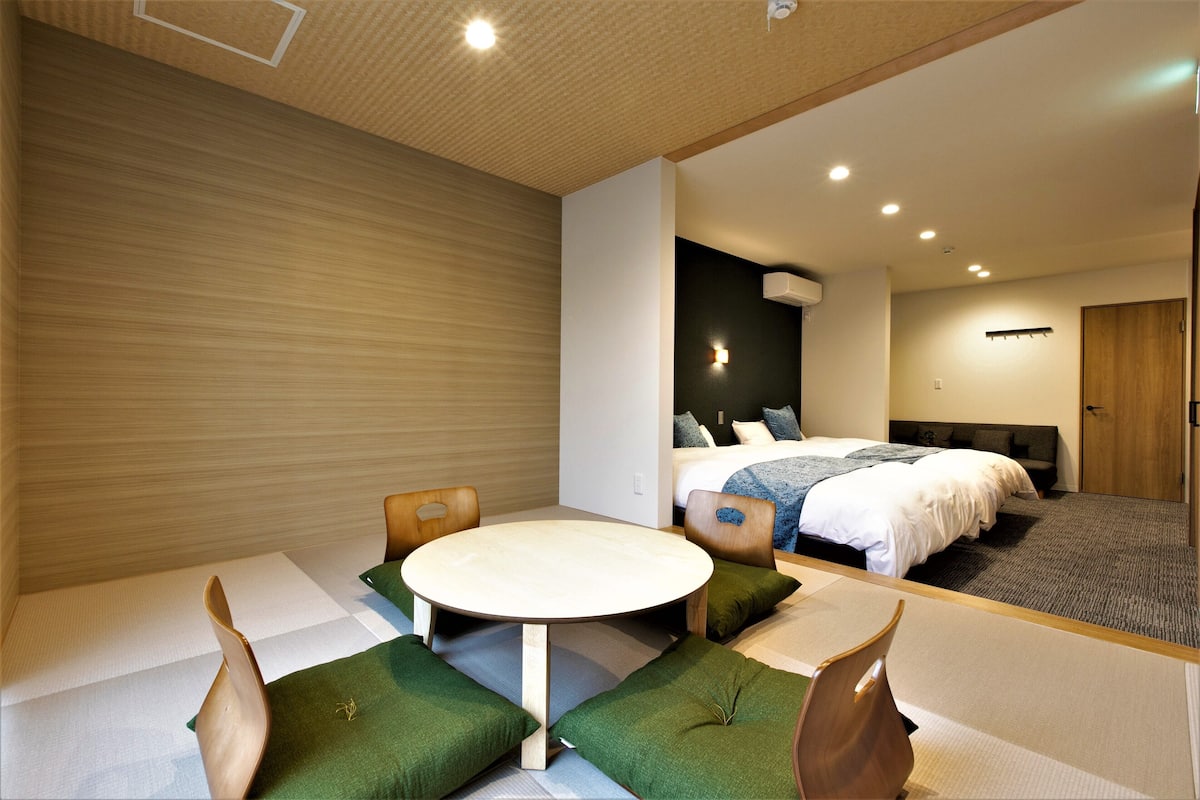 Una habitación de hotel moderna con una mesa redonda, sillas con cojines verdes, una cama con colcha blanca y un cabecero oscuro.
