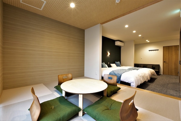 Una habitación de hotel moderna con una mesa redonda, sillas con cojines verdes, una cama con colcha blanca y un cabecero oscuro.